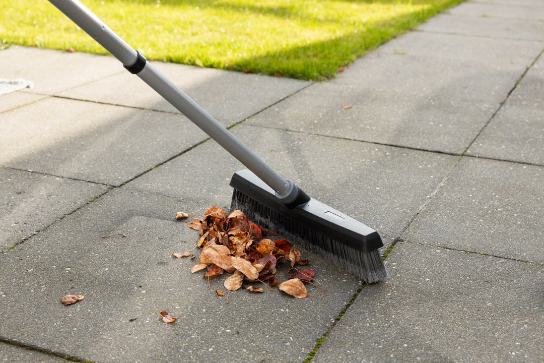 A broom sweeping up a pile of dry leaves on an outdoor patio with grass visible in the background