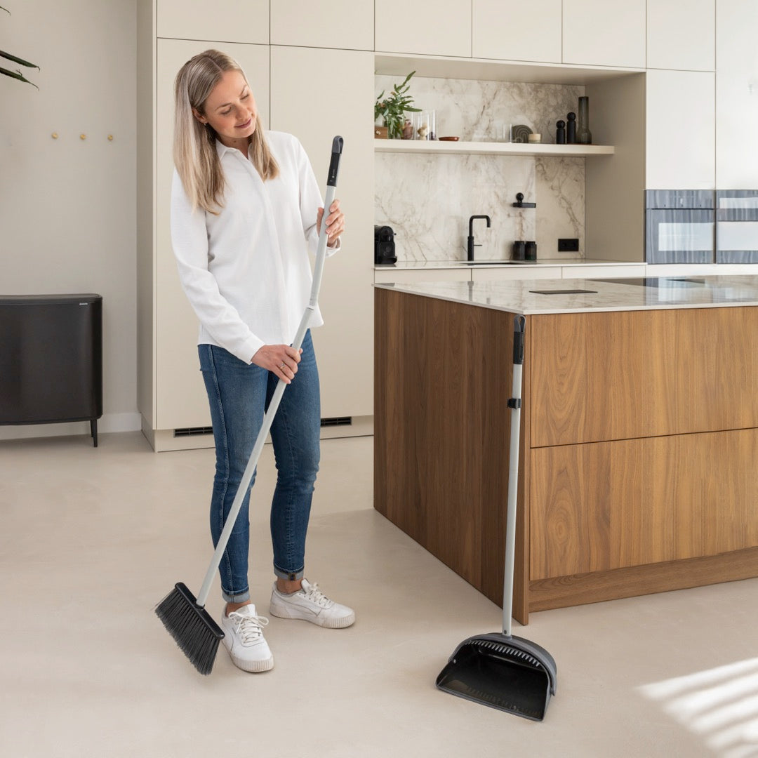 Woman holding a broom while standing in a modern kitchen, with a dustpan placed next to the kitchen island