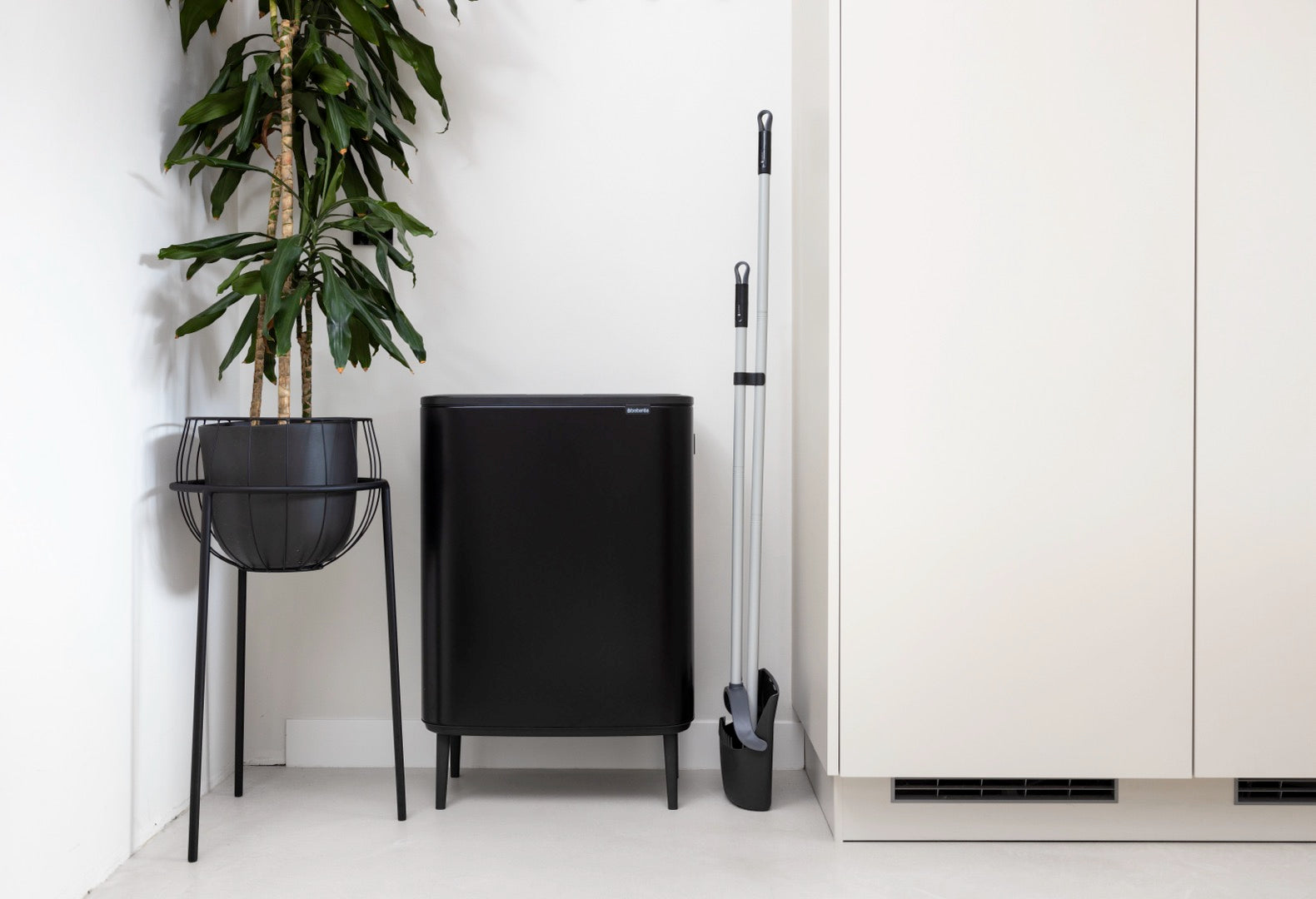 Broom and dustpan set stored neatly next to a black trash bin and a plant in a modern interior