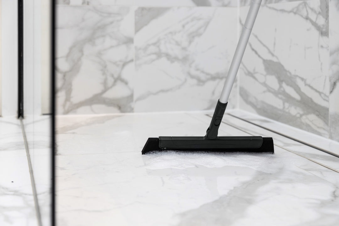 Close-up of a floor squeegee cleaning soapy water from a marble shower floor