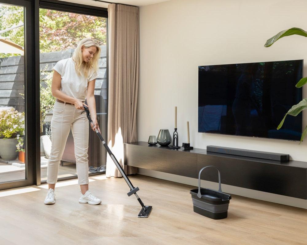 Woman using a mop set on a wooden floor in a modern living room, with a black bucket on the floor and large windows in the background