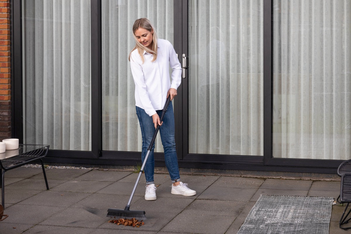 Woman sweeping leaves on a patio with a broom, standing near a glass door